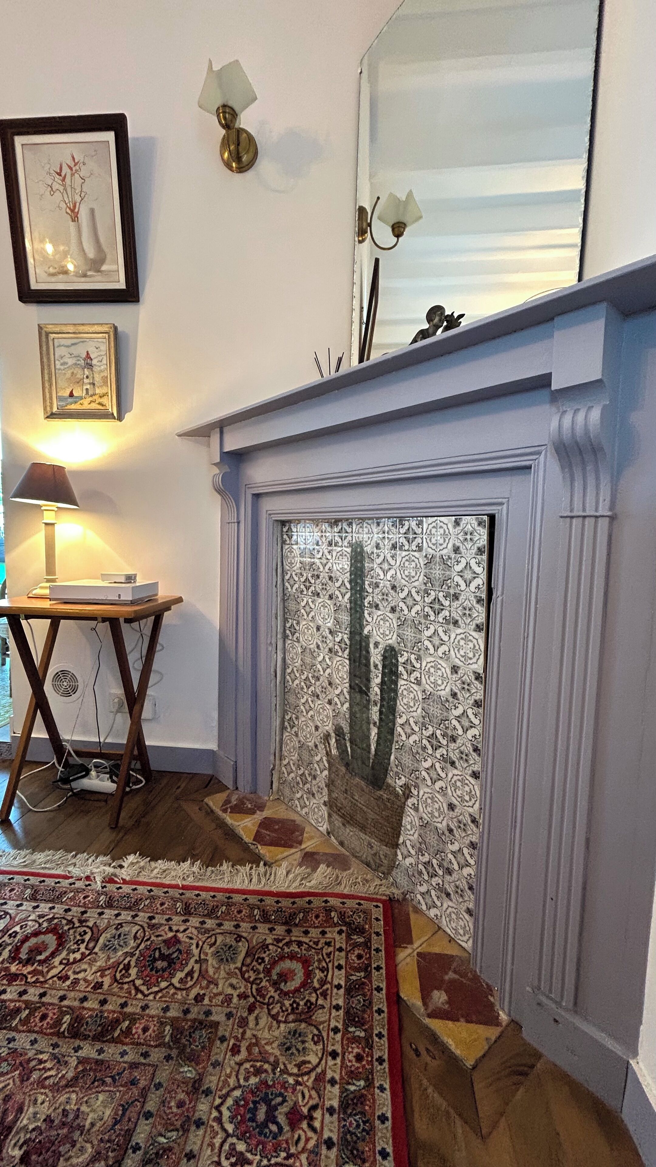 Un salon avec une cheminée, un tapis à motifs, une petite table avec une lampe et des œuvres d’art encadrées accrochées au mur.