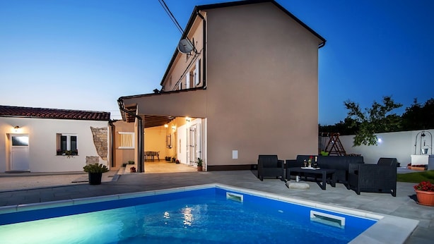 Una casa de dos pisos con piscina, mobiliario exterior y un patio cubierto.