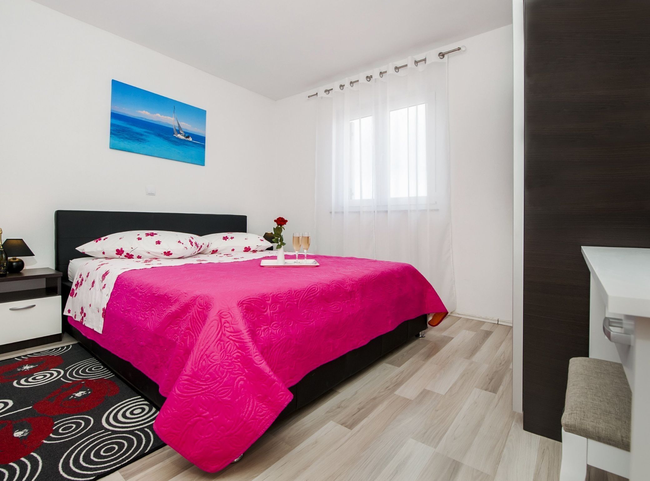 A modern bedroom with a bed, a nightstand, a desk, a window with curtains, and a painting on the wall.