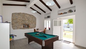 Mesa de billar en una habitación con pared de piedra, vigas de madera, dardo y vistas a una piscina exterior.