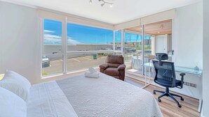 Quarto de hotel moderno com uma cama grande, uma escrivaninha com cadeira e vista para um estacionamento através de amplas janelas.