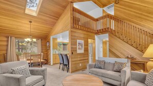 A spacious living area with a high ceiling, wooden walls, and a staircase leading to an upper level. There are two sofas and a round coffee table in the center. - Recently updated, great location, private hot tub! (Sunriver)
