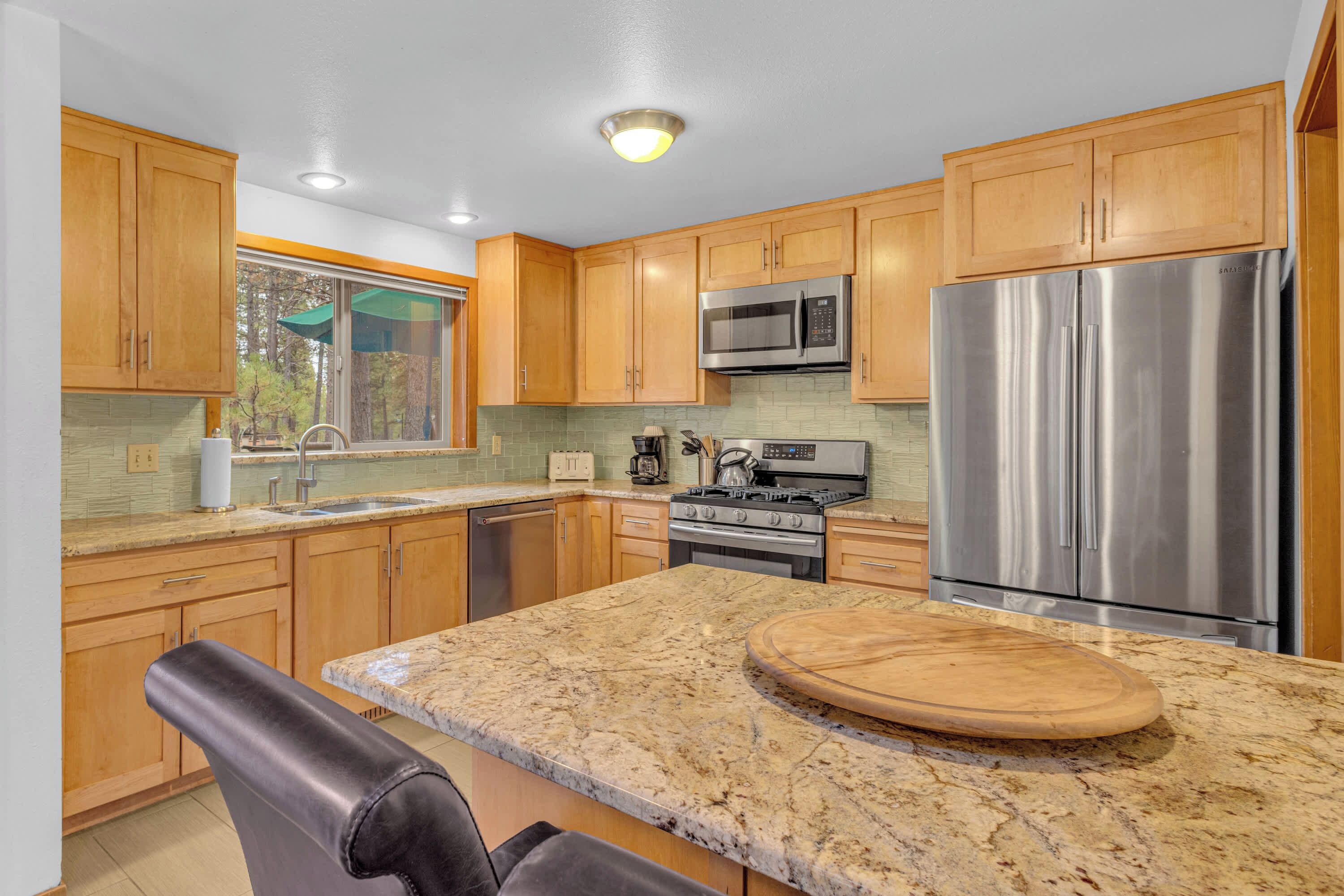A modern kitchen with wooden cabinets, stainless steel appliances, a granite countertop, and a window overlooking a forest.