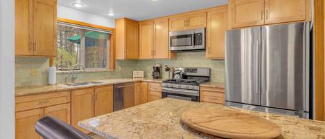 A modern kitchen with wooden cabinets, stainless steel appliances, a granite countertop, and a window overlooking a forest.