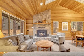 A cozy living room with a stone fireplace, wooden ceiling, and large windows.