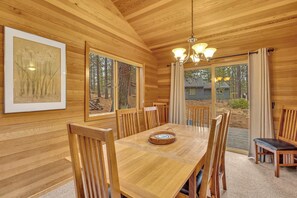 A wooden interior with a dining table, chairs, a chandelier, and a large window with a view of trees.