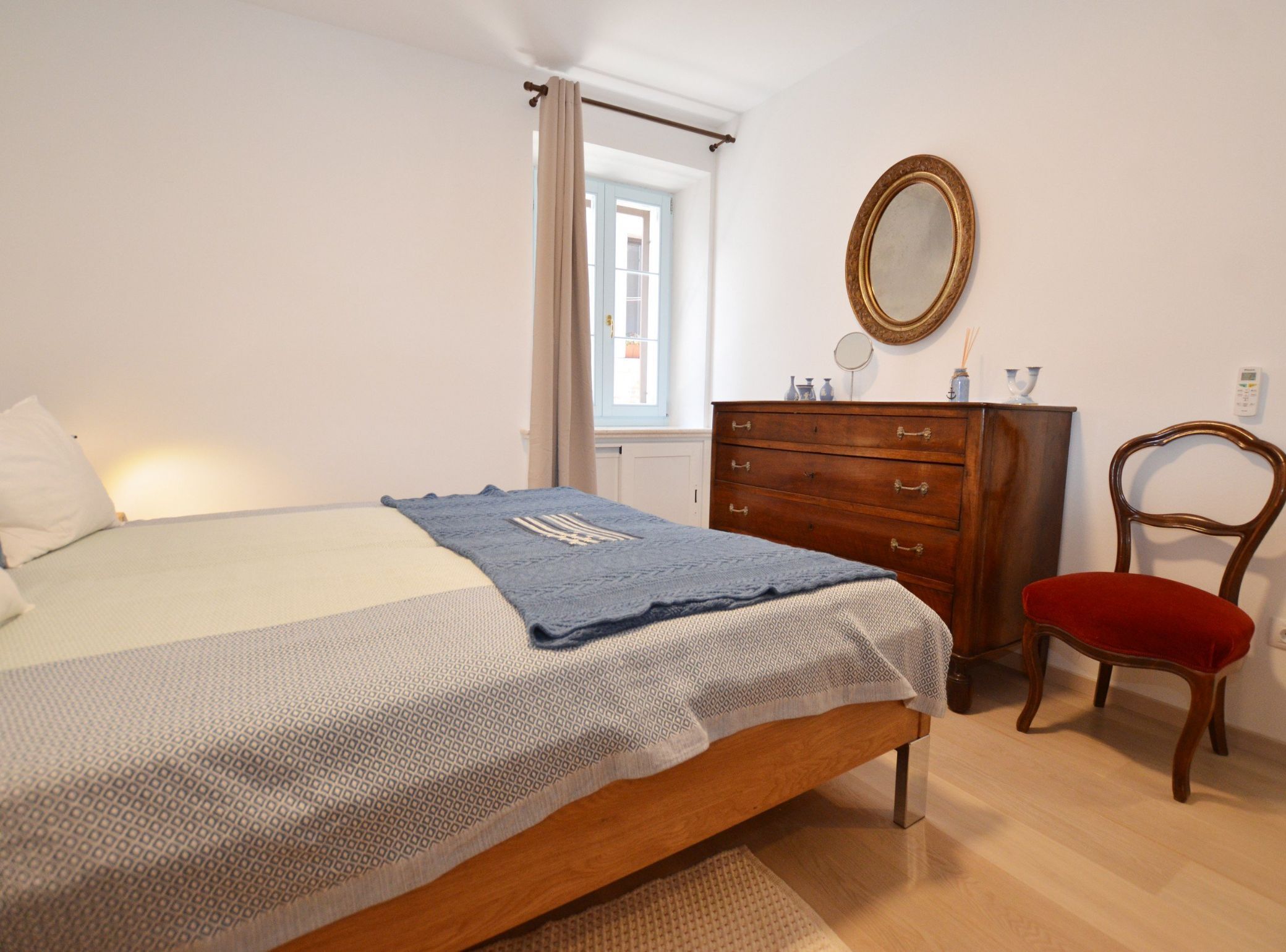 A bedroom with a bed, a wooden dresser, a chair, a mirror, and a window with curtains.