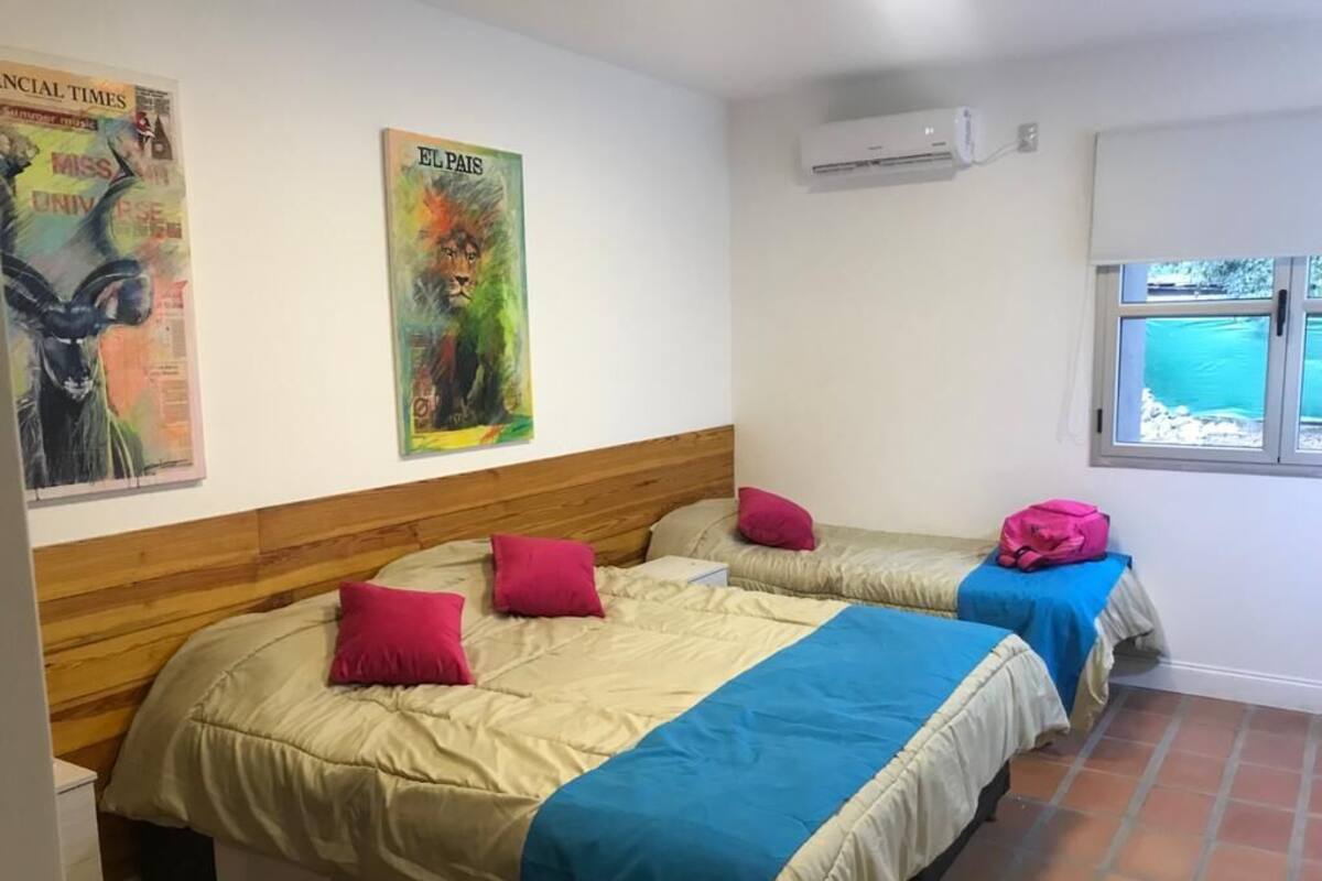 A room with two beds, each with a wooden headboard, a window with a view of greenery, and an air conditioning unit on the wall.