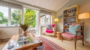 A cozy living room with a wooden coffee table, a patterned rug, a white armchair, a red and blue patterned chair, a bookshelf, and a view of greenery outside. - Corner Cottage, Brancaster, Norfolk (Brancaster)