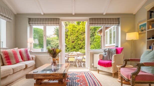 A cozy living room with a sofa, armchair, and coffee table, a dining area with chairs, and a view of greenery through large windows. - Corner Cottage, Brancaster, Norfolk (Brancaster)