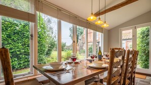 A dining area with a wooden table and chairs, large windows with a view of greenery and a brick building, and hanging pendant lights.