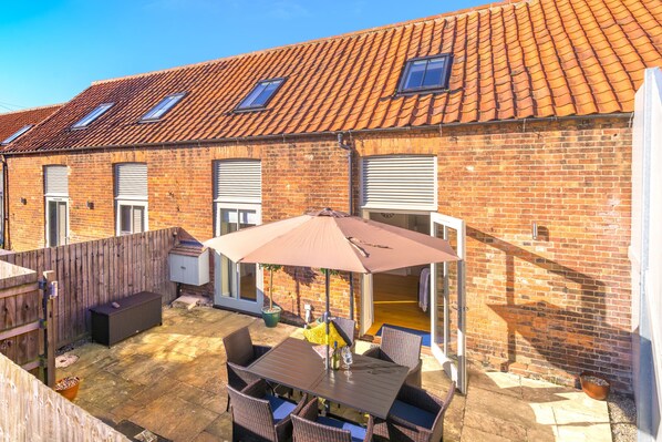 Un espace patio avec une table et des chaises, un grand parasol et un bâtiment en briques surmonté d’un toit en tuiles.
