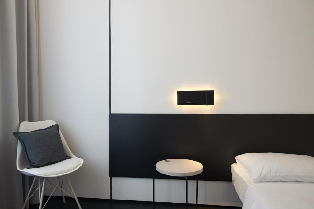 A modern hotel room with a bed, a small round table, a chair, and a wall-mounted light fixture.