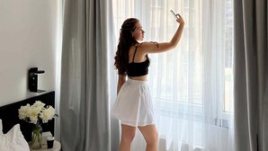 A hotel room with a bed, a bedside table with a vase of flowers, and a window with sheer curtains.
