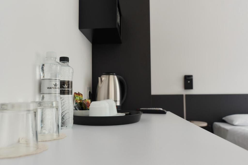 A modern hotel room with a white table featuring water bottles, glasses, a coffee pot, and a remote control.
