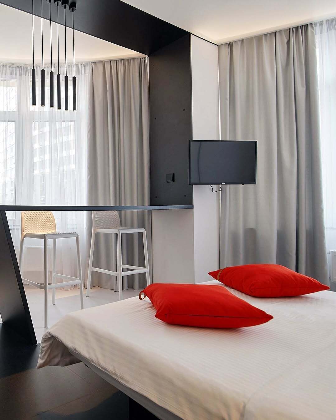 A modern hotel room with a bed, two red pillows, a TV, and a balcony with two chairs.
