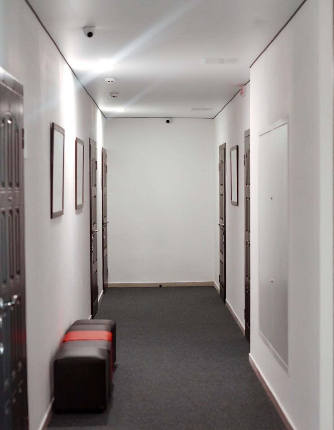 A narrow hallway with lockers on both sides, a grey carpet, and a security camera on the ceiling.