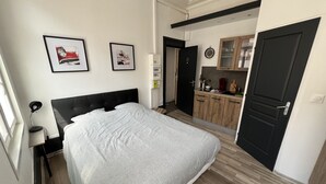 Una camera da letto con un letto, un comodino, una scrivania, una sedia, una finestra e un ingresso che dà su un angolo cottura.