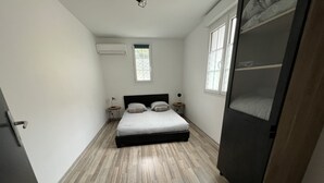 A bedroom with a bed, a nightstand, a lamp, a window, and an air conditioning unit. - Côté Jardin AMIENS 4-6 P (Amiens)