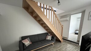 A modern living room with a grey sofa, wooden staircase, and a small round table with a remote control. - Côté Jardin AMIENS 4-6 P (Amiens)