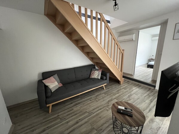 Un salon moderne avec un canapé gris, un escalier en bois et une petite table ronde sur laquelle se trouve une télécommande.