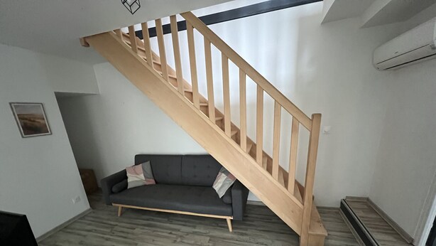 A modern living room with a wooden staircase, a grey sofa, and a wooden coffee table. - Côté Jardin AMIENS 4-6 P (Amiens)