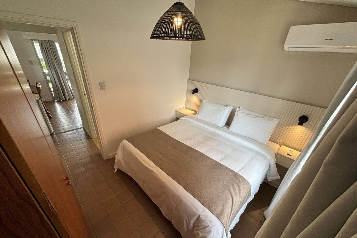 A hotel room with a bed, bedside lamps, a ceiling light, and an air conditioning unit.