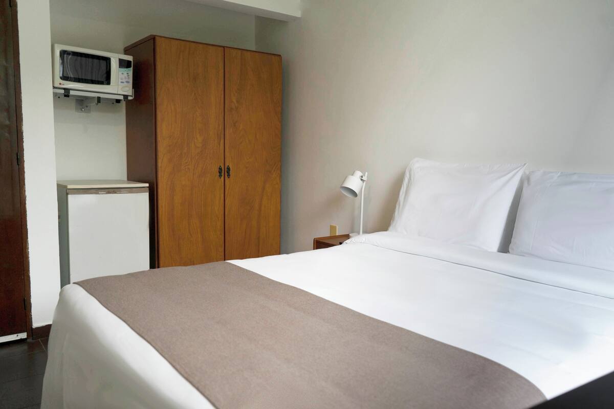 A hotel room with a bed, a wardrobe, a small bedside table with a lamp, and a microwave above the wardrobe.