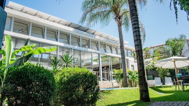 A modern hotel with a glass facade, a pool area, and lush greenery including palm trees and tropical plants.