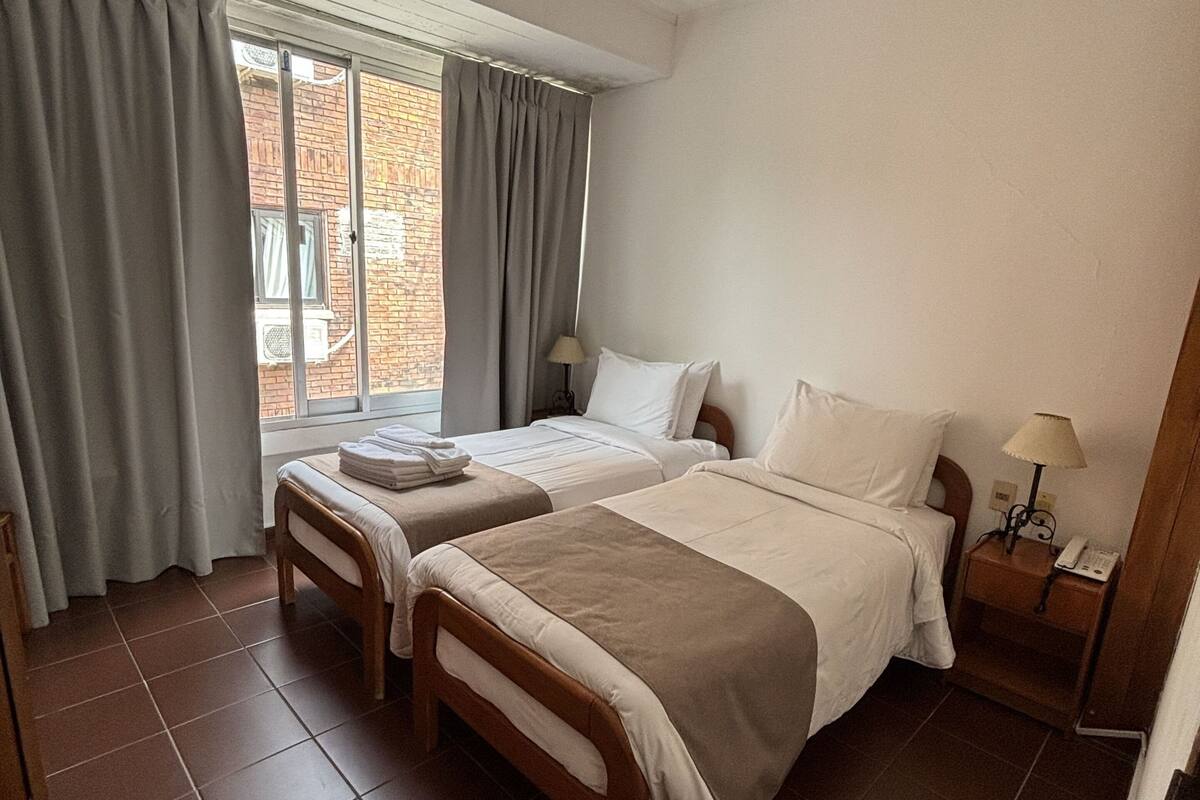 A hotel room with two beds, a window with curtains, a bedside table with a lamp, and a view of the outside.