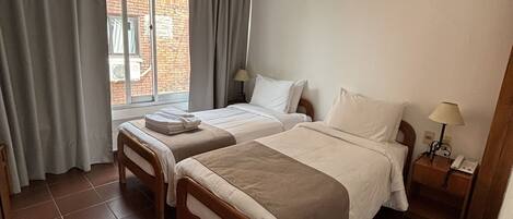 A hotel room with two beds, a window with curtains, a bedside table with a lamp, and a view of the outside.