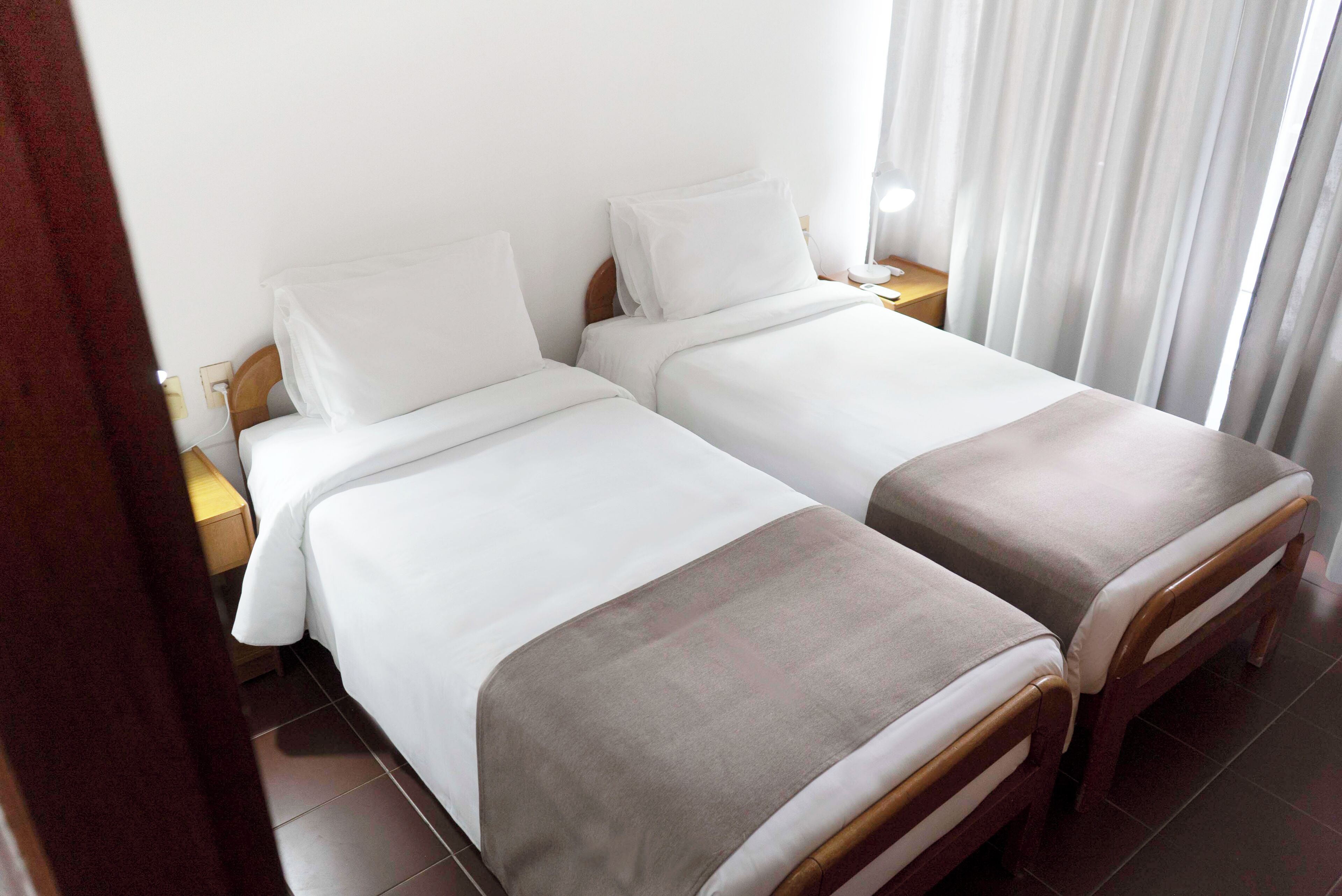 A hotel room with two single beds, a wooden nightstand, a lamp, and a window with curtains.