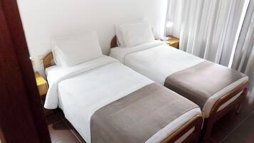 A hotel room with two single beds, a wooden nightstand, a lamp, and a window with curtains.