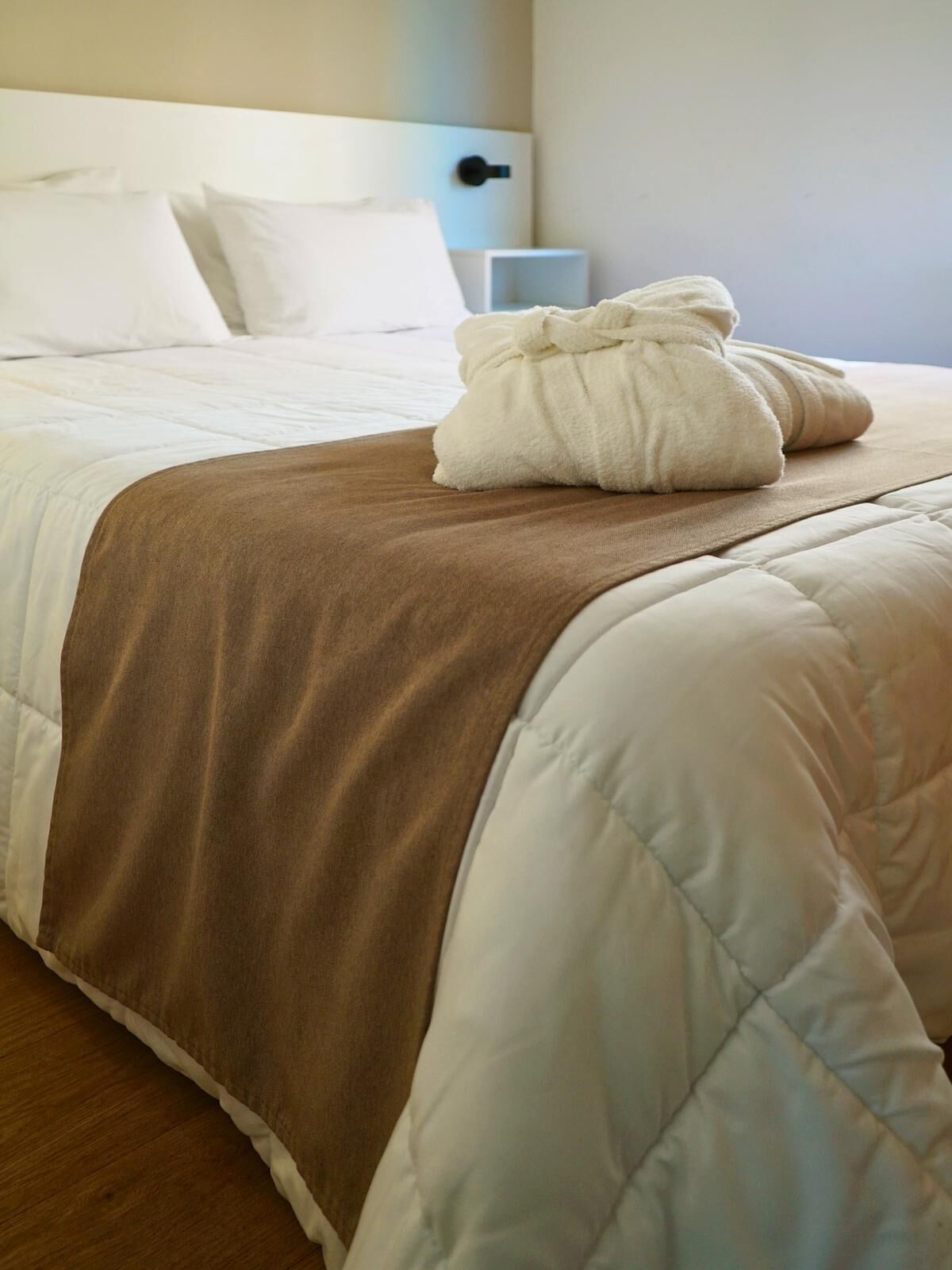 Una cama bien hecha con colcha marrón y blanca, una almohada blanca y una toalla doblada encima.