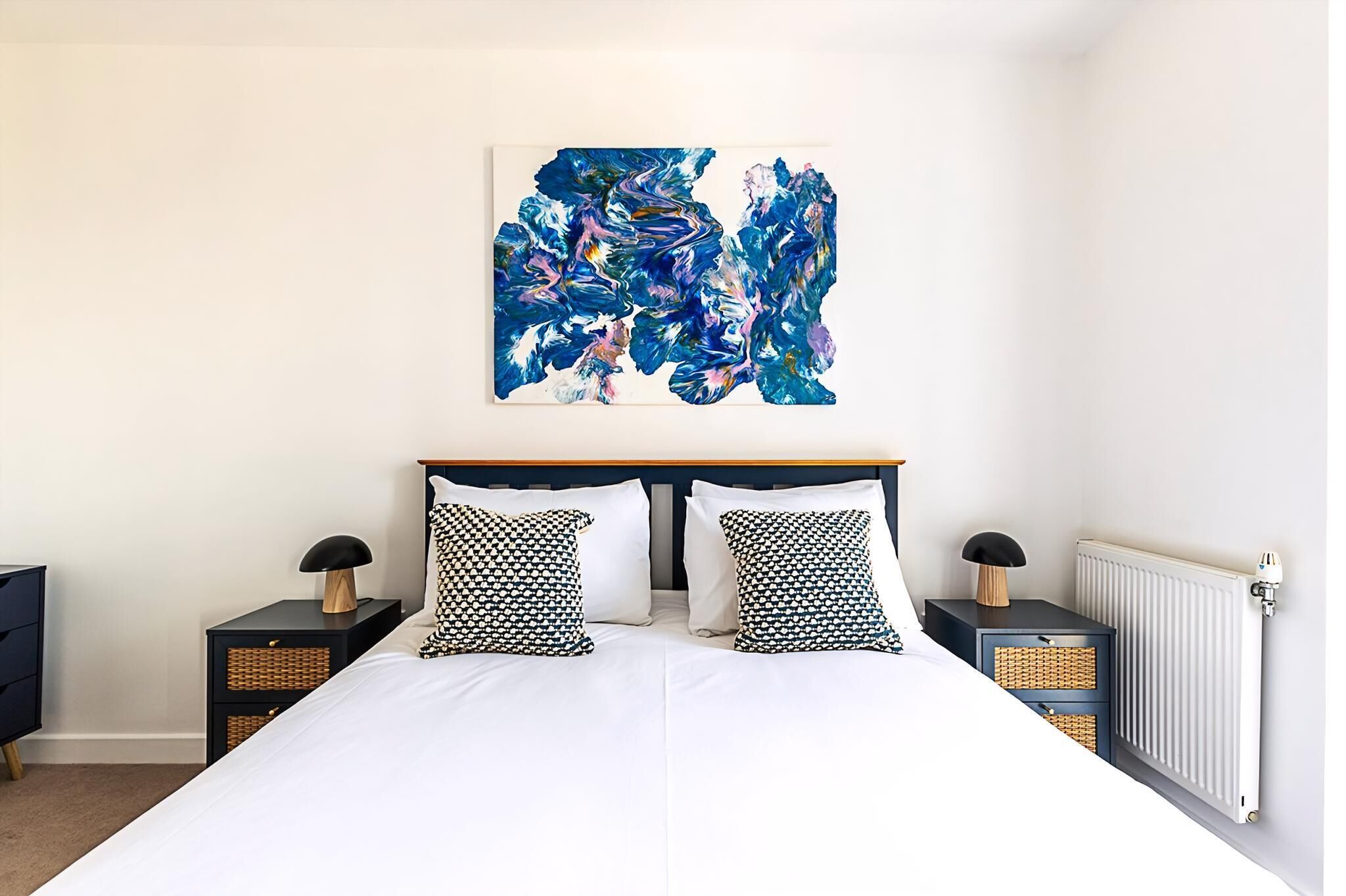 A bedroom with a large bed, two nightstands, a radiator, and a wall-mounted abstract painting.