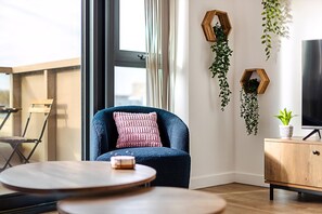 Ein modernes Wohnzimmer mit blauem Sessel, rundem Holztisch, Holzschrank und großem Fenster mit Vorhängen.