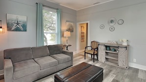 A living room with a grey sofa, a black ottoman, a wooden chair, and a small table with a lamp.