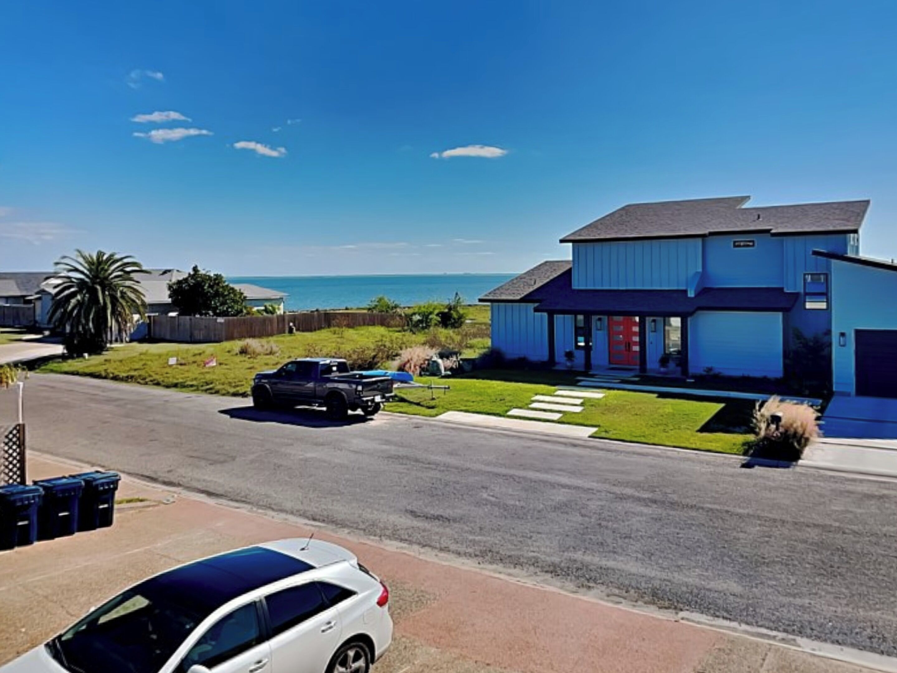 Een modern huis met twee verdiepingen, met een rode deur en een garage, gelegen aan het einde van een straat met vrij uitzicht op de oceaan.