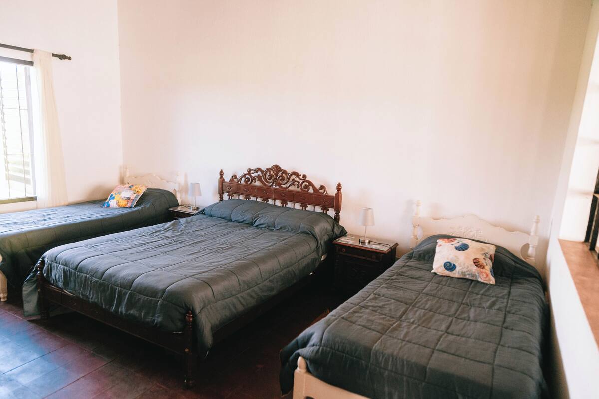 Quarto com três camas individuais, cada uma com cabeceira de madeira e mesa de cabeceira com luminária.