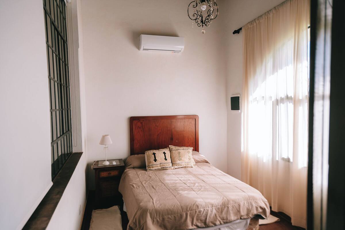 Quarto com cabeceira de madeira, cama com roupa de cama bege, mesa de cabeceira com abajur e unidade de ar condicionado na parede.