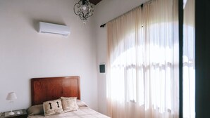 Een slaapkamer met een houten hoofdbord, een bed met beige beddengoed, een nachtkastje met een lamp en een raam met dunne gordijnen.