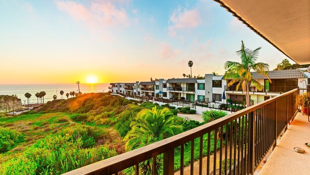 A hotel with multiple balconies overlooking a coastal area, featuring lush greenery and a view of the ocean at sunset.