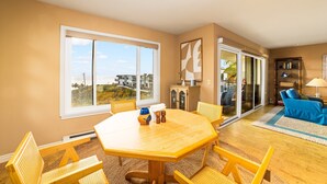 A spacious living room with a large wooden table, comfortable seating, a view through a sliding glass door, and a bookshelf.