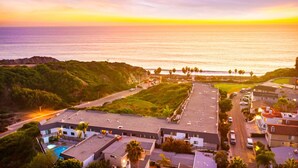 A coastal hotel with a swimming pool, parking area, and views of the ocean and hills.