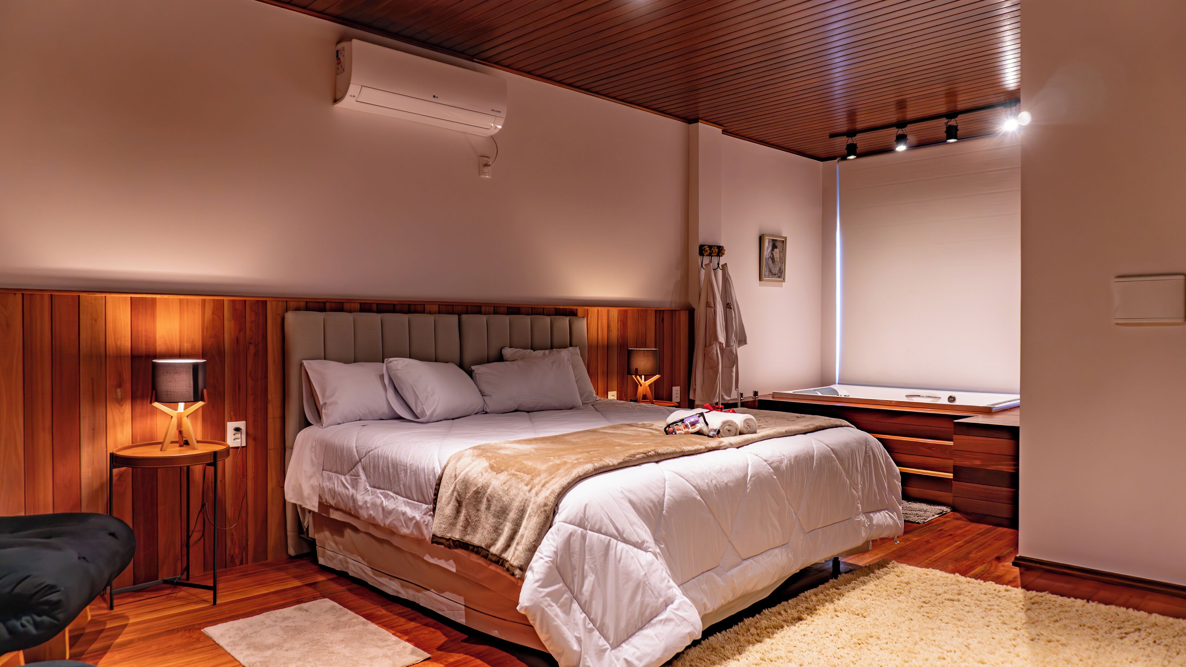Un dormitorio con cabecera de madera, una cama con ropa de cama blanca, una mesita de noche con lámpara, suelo de madera y un espejo grande en la pared.