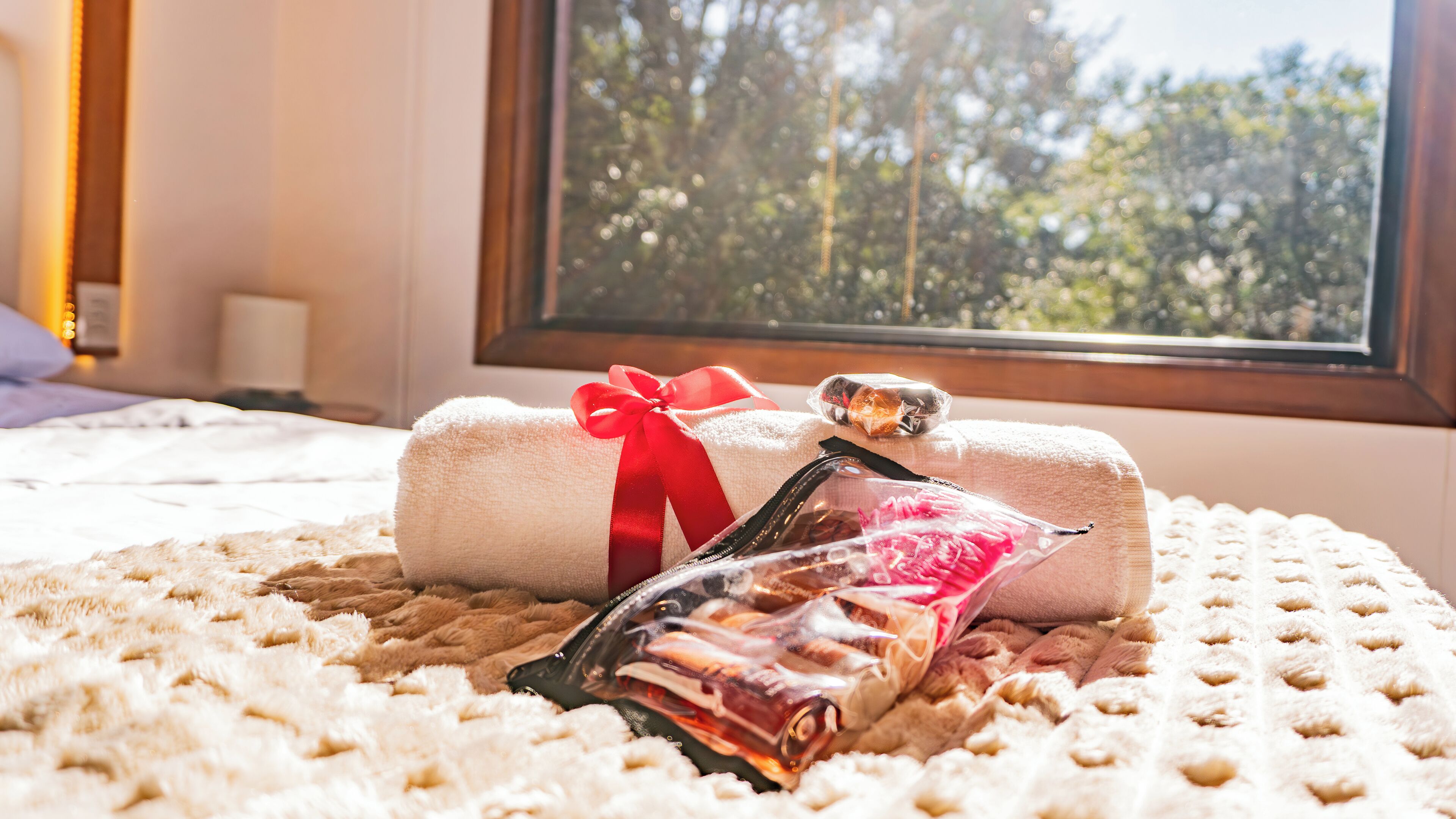 Ein ordentlich bezogenes Bett mit einem zusammengerollten Handtuch und einer Geschenktüte auf einer gemusterten Decke, mit Blick auf Bäume durch ein Fenster.