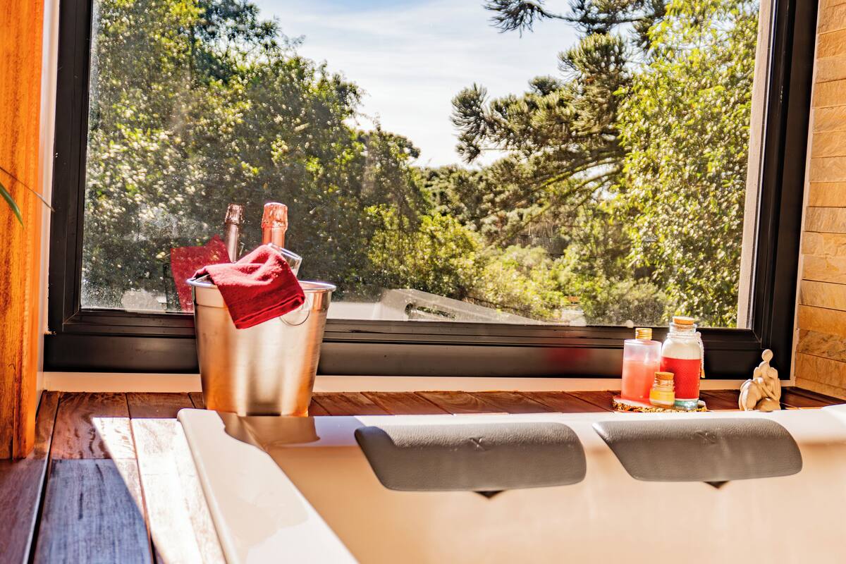 A bathtub with a view of trees through a window, a bucket with a towel, and two bottles on the side.