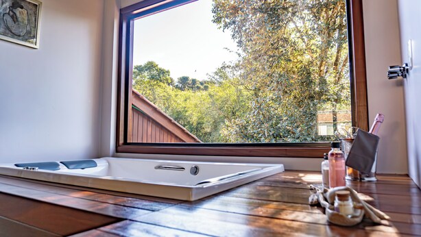 A room with a large window, a wooden floor, a white bathtub, and a small table with toiletries. - Invernada Blessmann  (Vespasiano Corrêa)