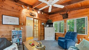 A cozy cabin interior with wooden walls, a ceiling fan, a bookshelf, a television, and a large window with a view of trees. - SHORT WALK TO A LARGE SANDY BEACH (FRYE ISLAND)