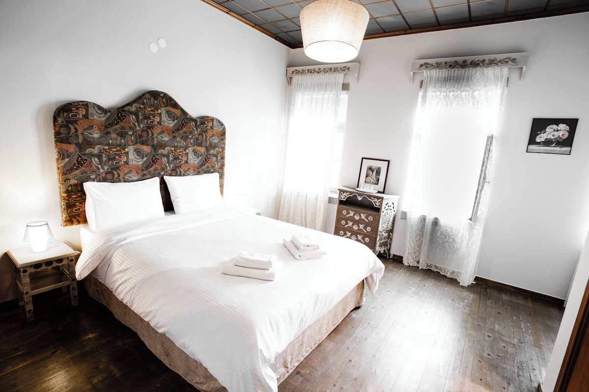 Een slaapkamer met een groot bed, een houten hoofdbord met een gedessineerd patroon, een nachtkastje met een lamp en een dressoir met een fotolijst.